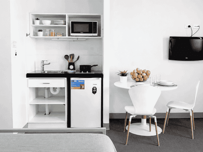 Compact studio with kitchenette, white dining table, chairs, wall-mounted TV, and modern minimalist design.