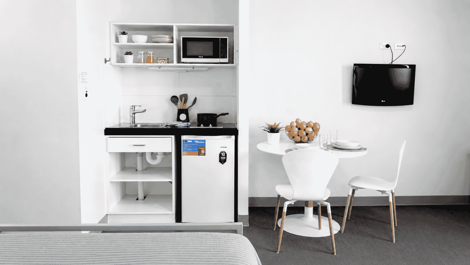 Compact studio with kitchenette, white dining table, chairs, wall-mounted TV, and modern minimalist design.