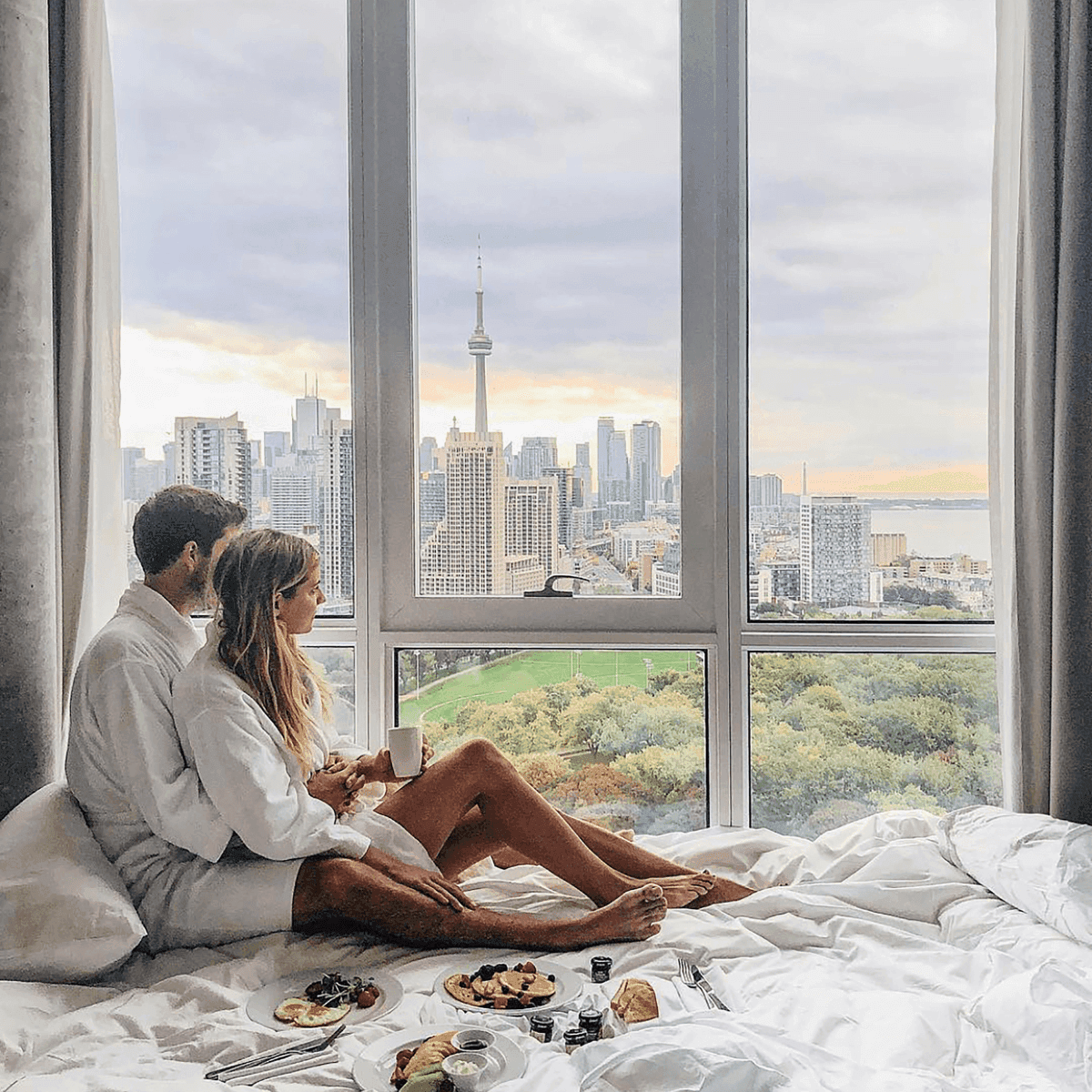 Couple sharing breakfast plates on the bed by a window, under panoramic views of the skyline at Hotel X Toronto