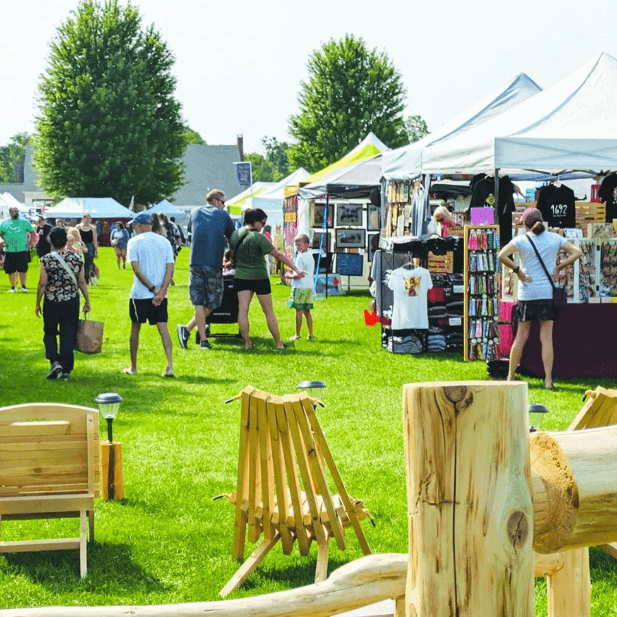 Crowded outdoor crafting fair with people browsing vendor stalls, children playing, and wooden chairs on grassy field.