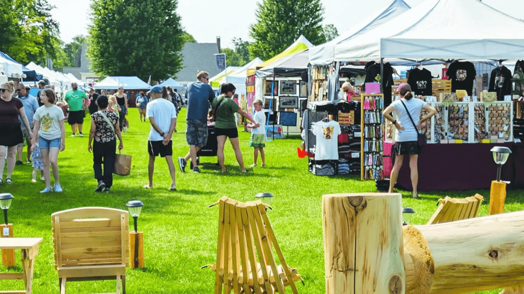 Crowded outdoor crafting fair with people browsing vendor stalls, children playing, and wooden chairs on grassy field.