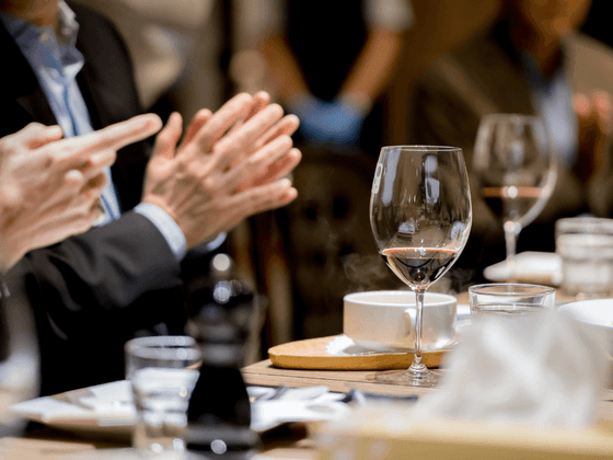 Closs-up of a glass of red wine on a dining table surrounded by hands applauding at Hotel des Innovations