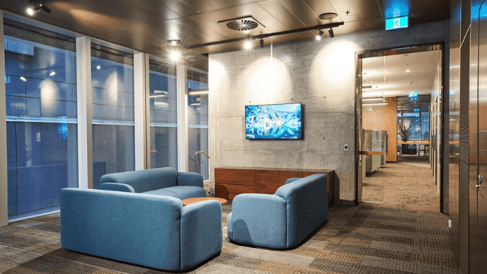 Modern lounge with blue couches, flat-screen TV on wall, large windows, and hallway.