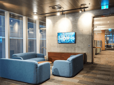 Modern lounge with blue couches, flat-screen TV on wall, large windows, and hallway.