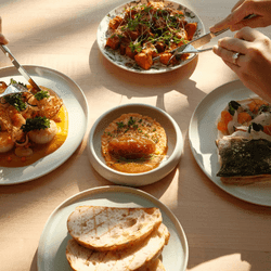 Hands hold cutlery over four plates of varied dishes on a table, with one plate containing sliced bread.