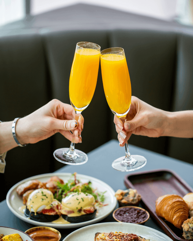 Mimosas raised in a toast by a table of eggs benedict, croissants and preserves at Hotel X Toronto