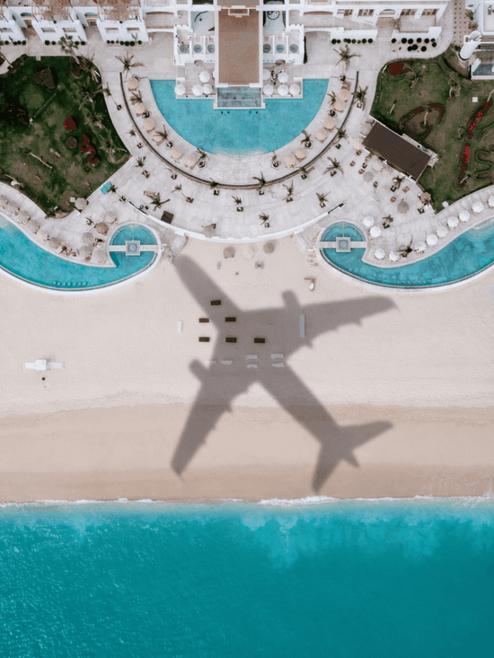 Aerial view of airplane shadow over beachfront resort with curved pools, palm trees, and white sand beach.