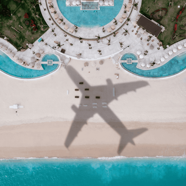 Vista aérea de resort con piscinas turquesas, playa y sombra de avión sobre la arena blanca