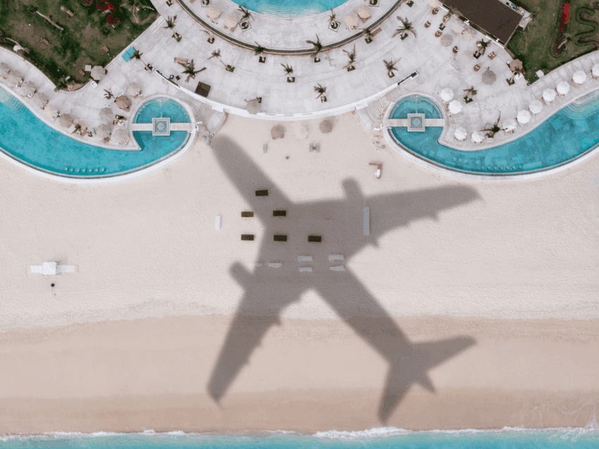 Aerial view of airplane shadow over beachfront resort with curved pools, palm trees, and white sand beach.