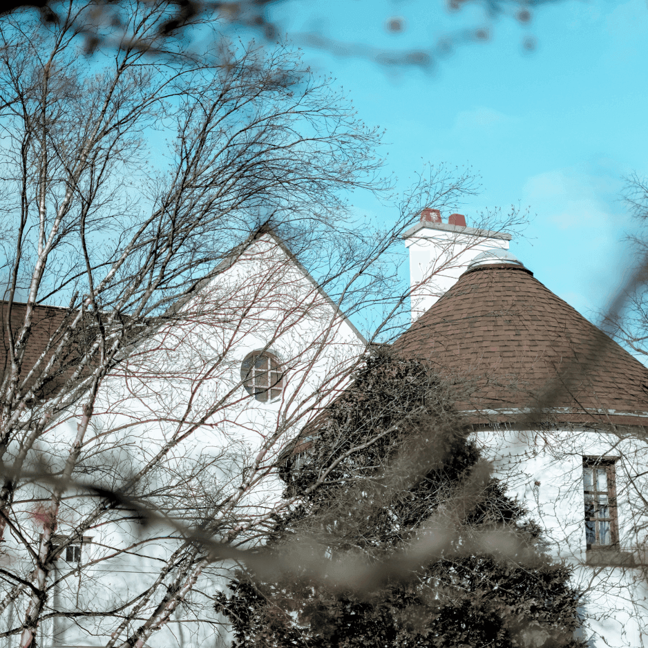 Conical turret roof and white gable of La Tourelle Inn & Spa, framed by bare branches near a chimney under blue sky