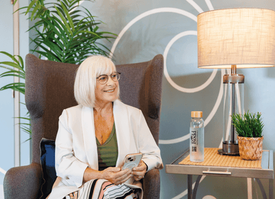 Smiling senior woman with smartphone relaxing on an armchair beside a lamp and branded water bottle inside Rodd Moncton