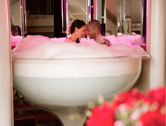 A couple sharing a romantic moment in a bubble-filled bathtub, surrounded by soft pink lighting at Cove Pocono Resorts