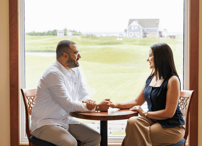 Couple seated at a round table by a large window overlooking the lush golf course at Rodd Crowbush Golf & Beach Resort