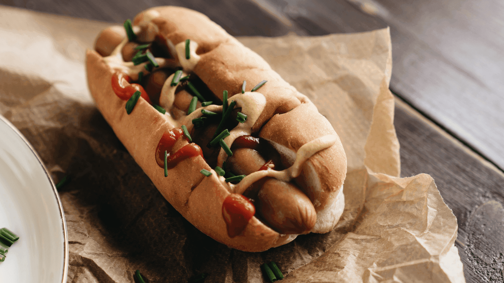Hot dog on bun with ketchup and chives on paper atop a wooden surface next to a plate.