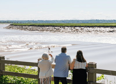 Three people leaning against a wooden fence watching a paddleboarder ride tidal bore waves outside the Rodd Moncton
