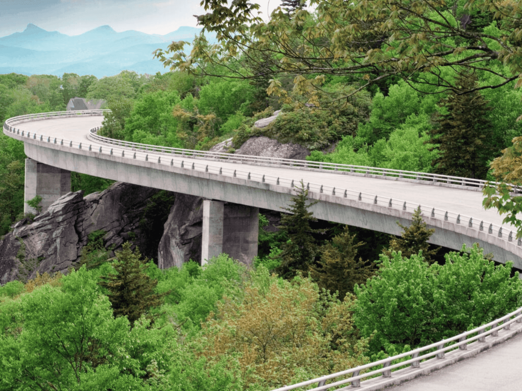 Elevated curved road bridge with safety rails over lush greenery and rocky terrain, with distant mountains.
