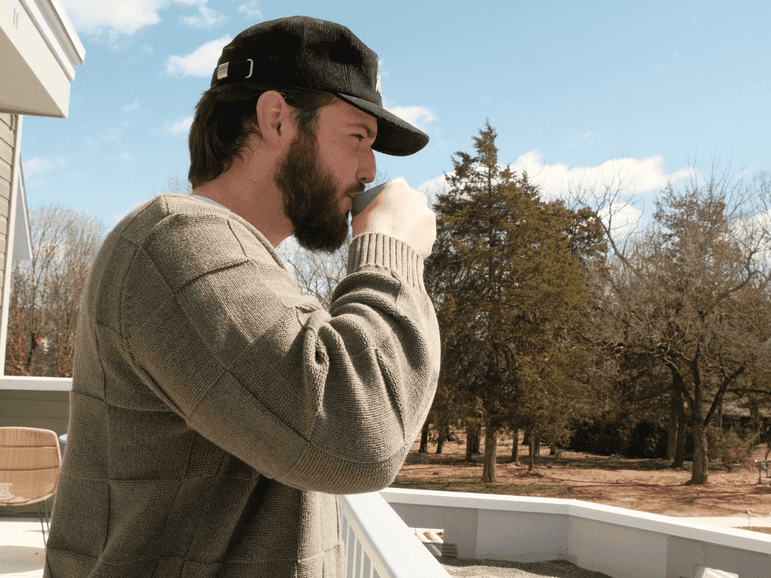 Man on a balcony wearing a hat and sweater, holding a cup, and facing a wooded area.