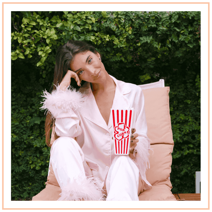 Woman in a soft pink, feathered pajama set relaxes outdoors, holding a popcorn tub at The Plymouth Hotel South Beach