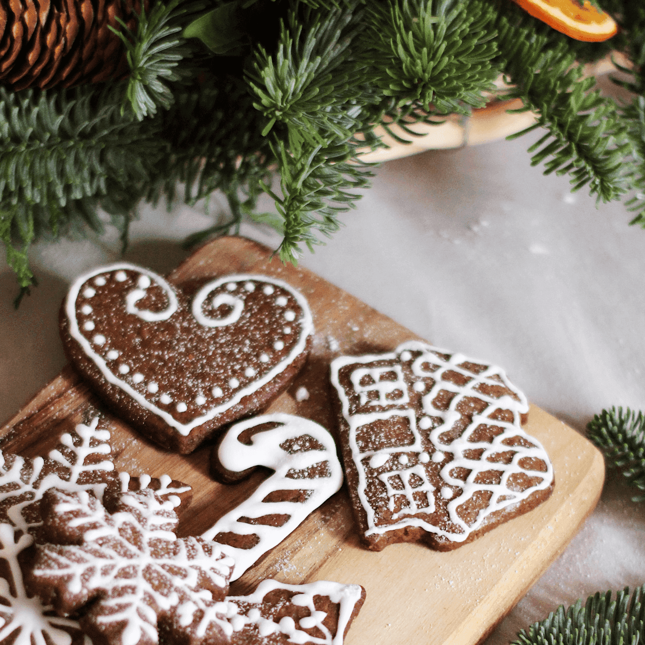 Wooden board with gingerbread cookies shaped like heart, candy cane, house, and snowflakes, surrounded by greenery.
