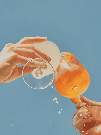 Two hands holding wine glasses filled with orange liquid, one with ice, against a blue background.