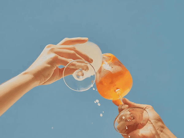 Two hands holding wine glasses filled with orange liquid, one with ice, against a blue background.