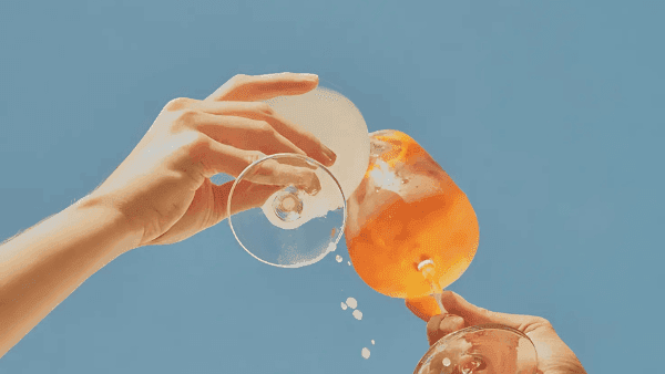 Two hands holding wine glasses filled with orange liquid, one with ice, against a blue background.