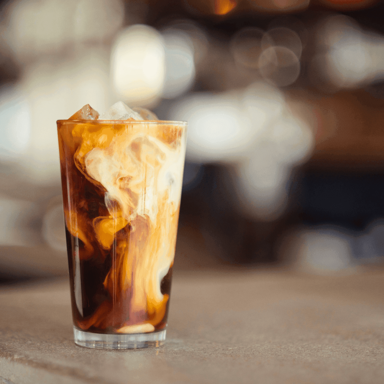 A glass of iced coffee with ice cubes on a table with a blurred background.