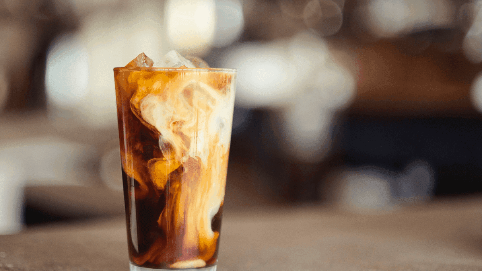 A glass of iced coffee with ice cubes on a table with a blurred background.
