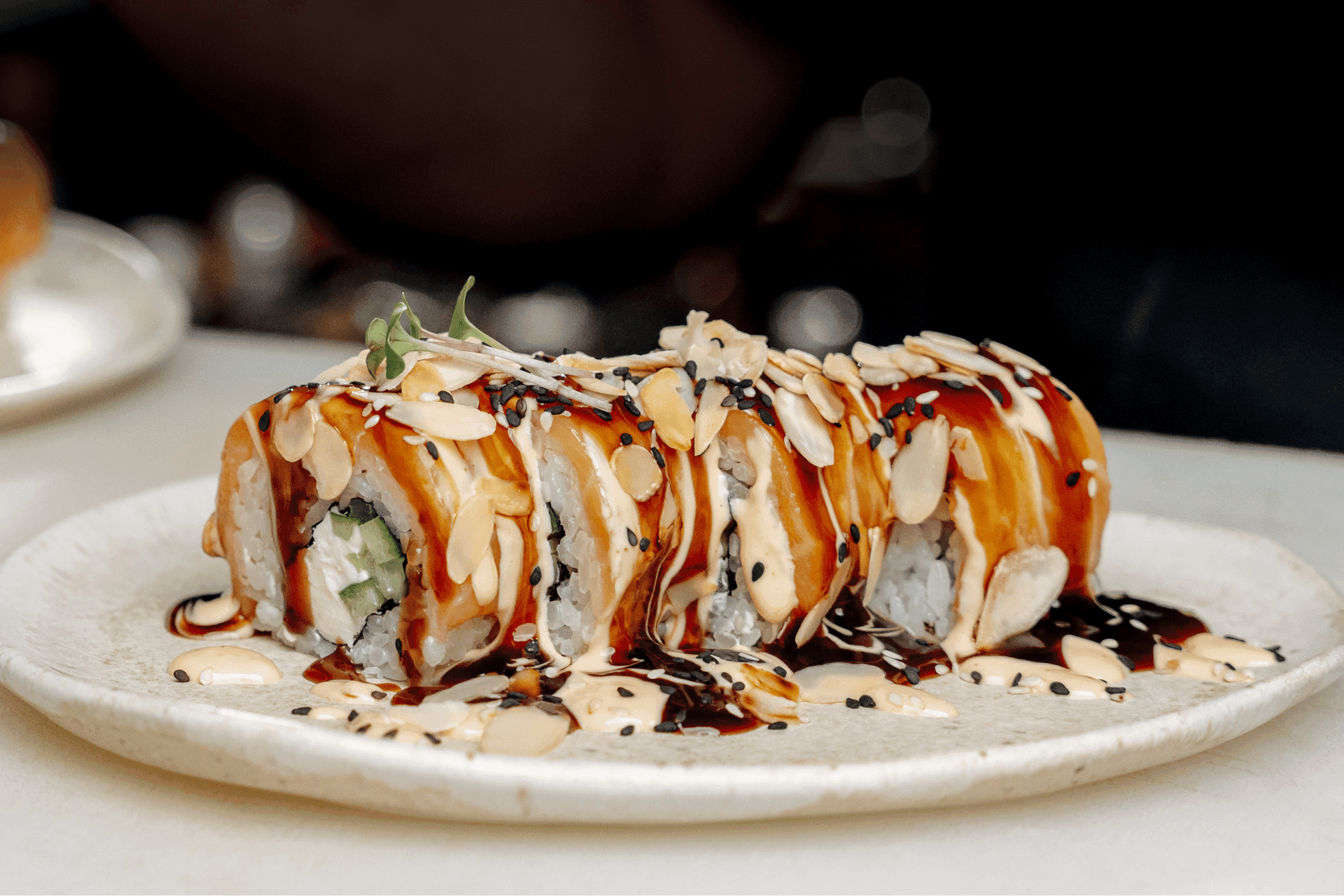 Photograph of a sushi roll plated on a white dish, showcasing a close-up view of the roll topped with sliced almonds, black sesame seeds, and drizzled with creamy and dark sauces. The sushi roll contains cucumber and other ingredients, with a garnish of microgreens on top, highlighting a visually appealing and flavorful presentation.