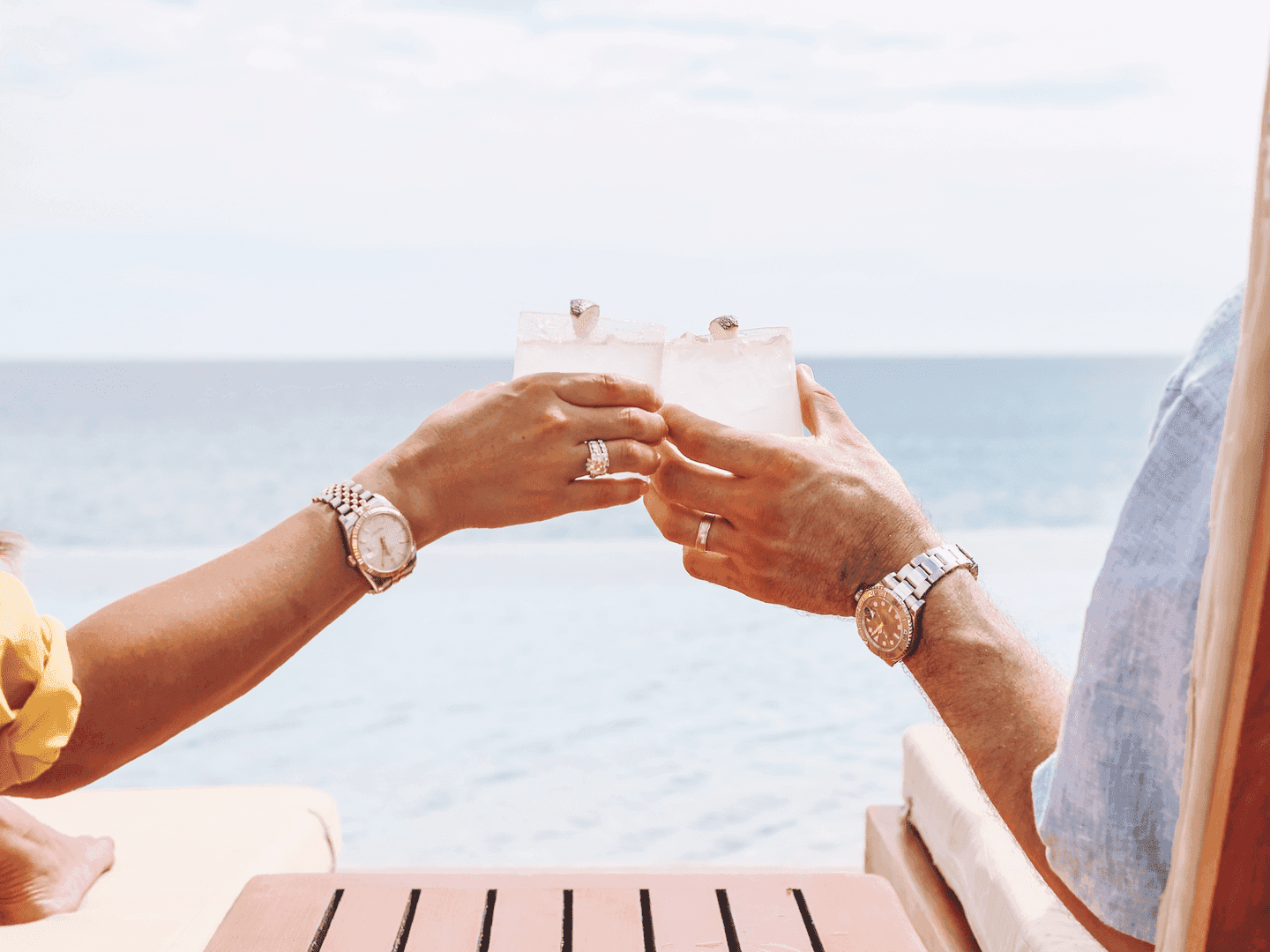 Two people toasting with tropical cocktails at a beachfront table overlooking the ocean under a sunny sky.