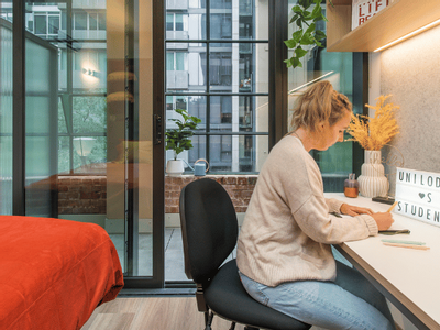 A woman is seated at a desk, working on a laptop, with a bed nearby in a modern apartment.