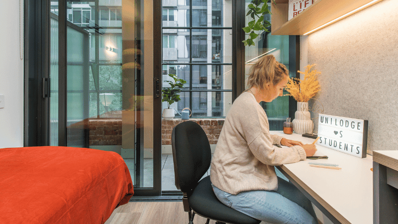 A woman is seated at a desk, working on a laptop, with a bed nearby in a modern apartment.