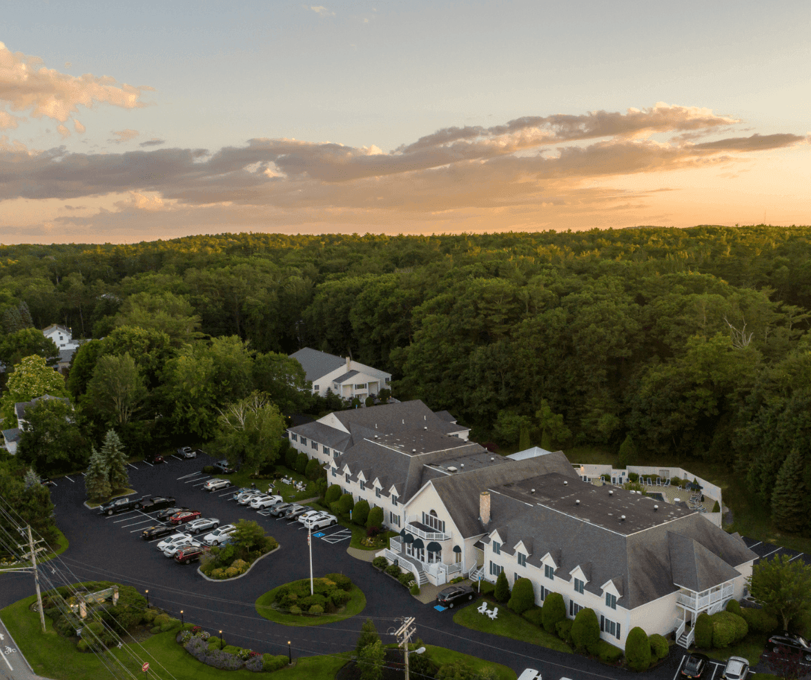 Aerial view of a white building with a gray roof surrounded by trees and a parking lot.