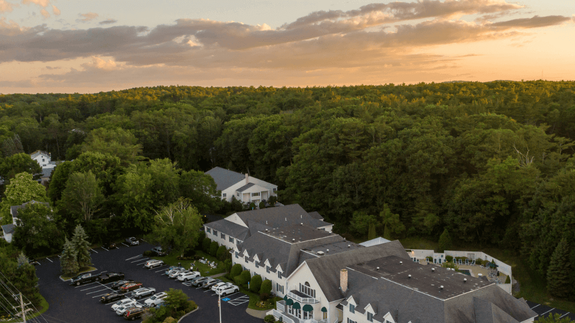 An aerial view of a white building with a gray roof surrounded by trees and a parking lot at sunset.