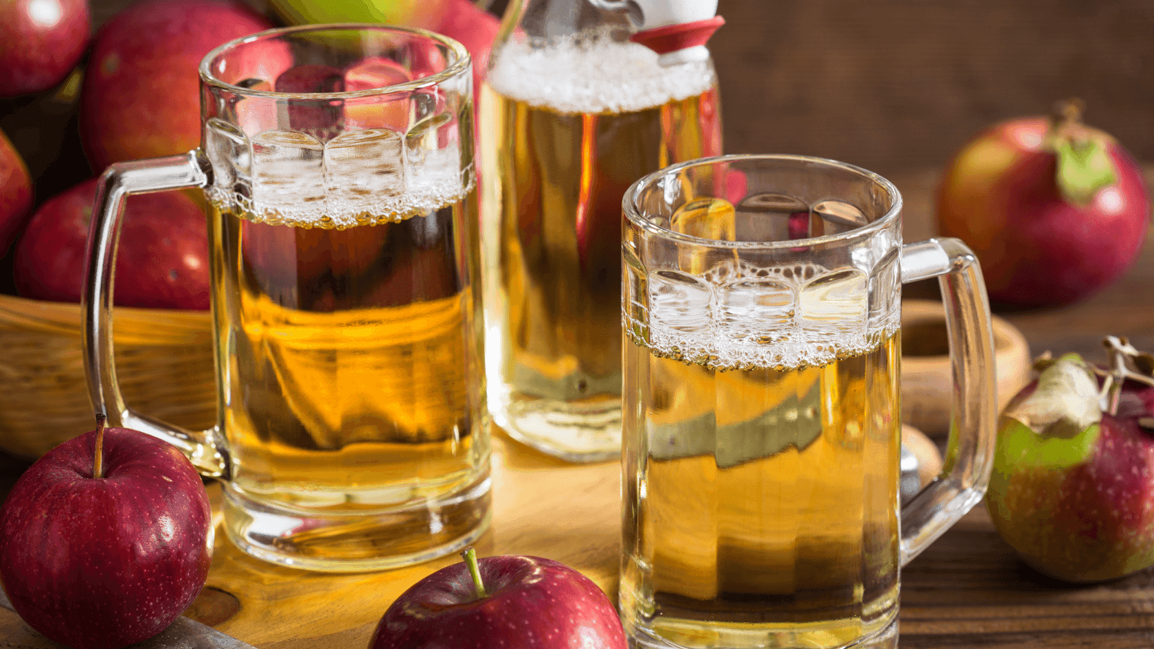 Two mugs of cider with apples and a bottle of cider on a wooden surface.