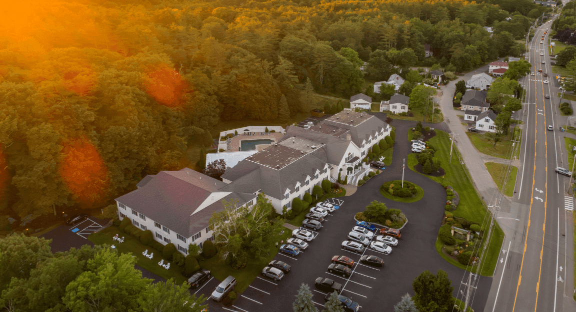 Aerial view of a white building with a gray roof surrounded by trees and a parking lot.
