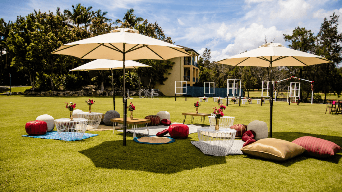Outdoor event setup with tables, chairs, umbrellas, and decorative pillows on a green lawn.