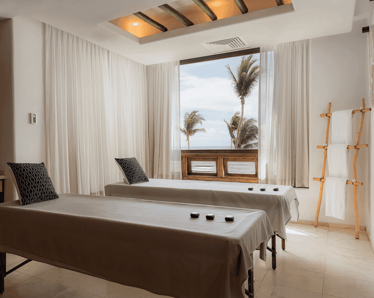 Massage treatment room with two tables, ocean view, palm trees, and wooden towel rack.