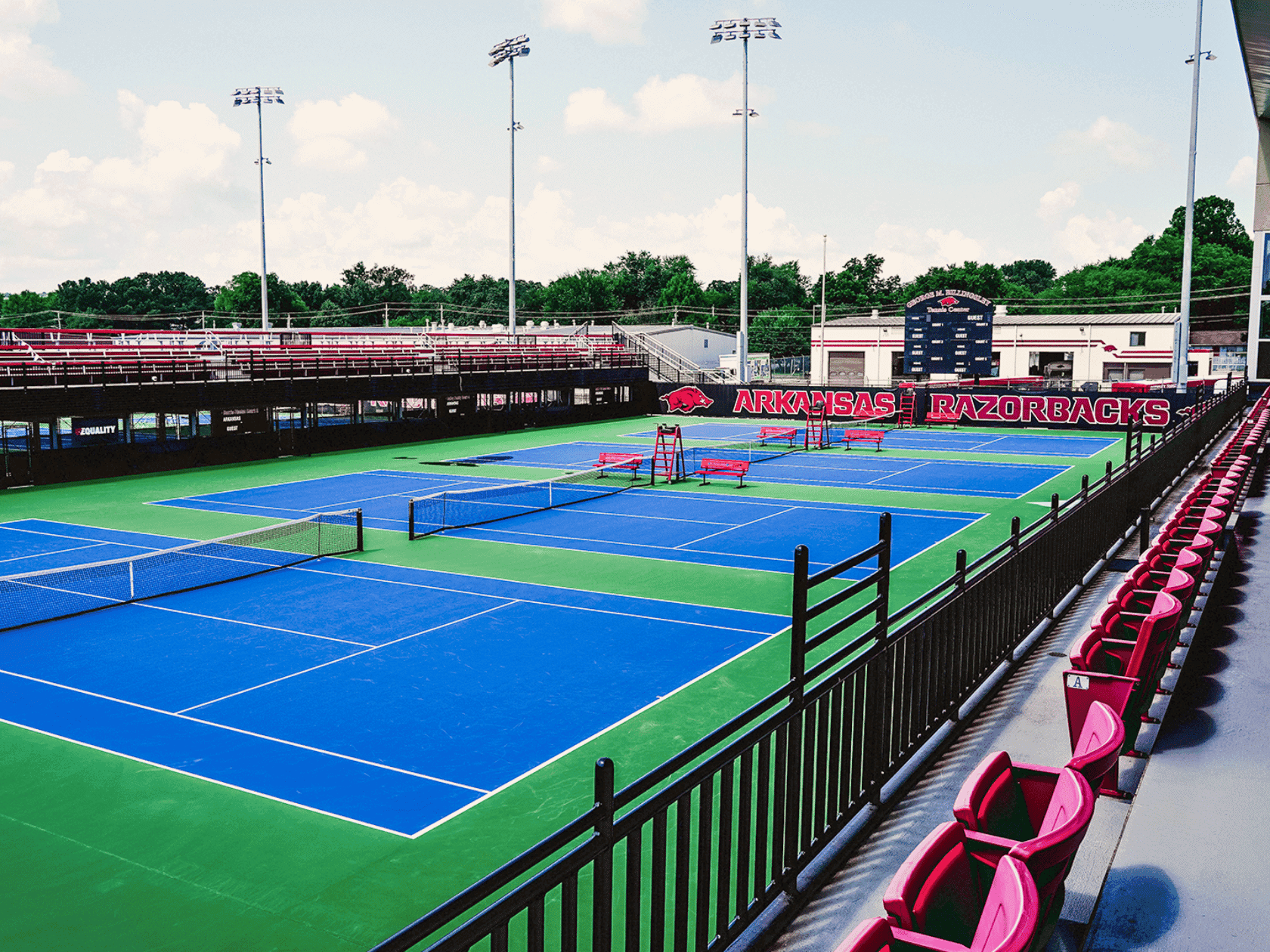 Arkansas Razorbacks tennis courts with blue and green surfaces, surrounded by black fencing and red spectator seating.