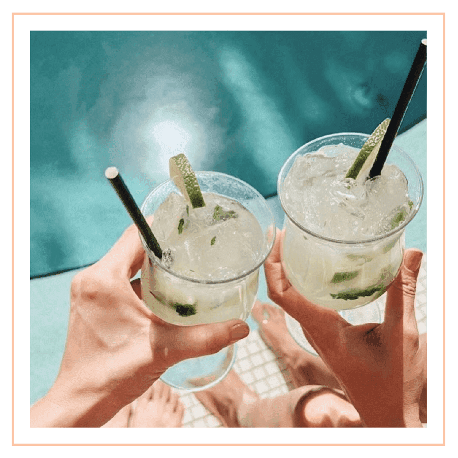 Two hands hold iced Caipirinha cocktails over a bright blue swimming pool on a sunny day at The Plymouth Hotel South Beach
