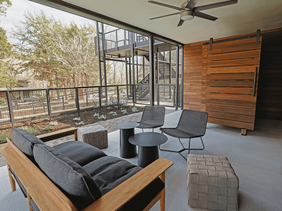Modern outdoor lounge with wooden furniture, two tables, two chairs, a large sliding door, and a balcony with railings.