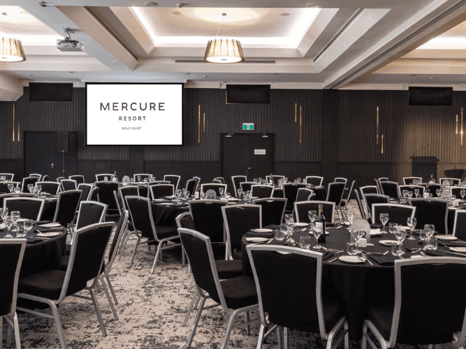Large banquet hall with rows of black tables and chairs, two large screens displaying