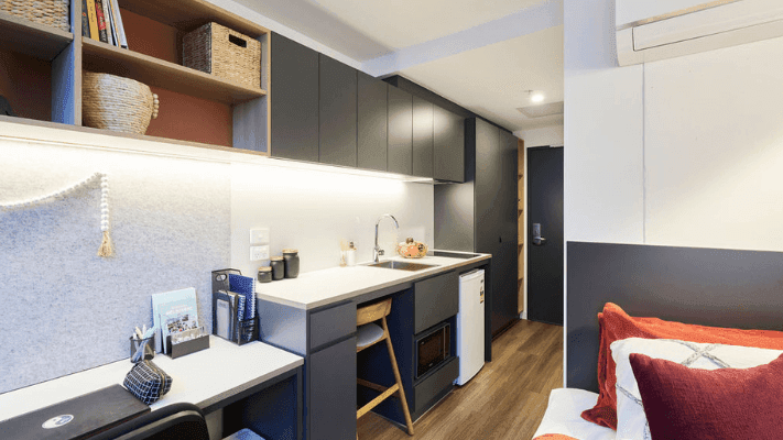 A compact studio apartment with a small kitchen, dining area, and a bed with red pillows.