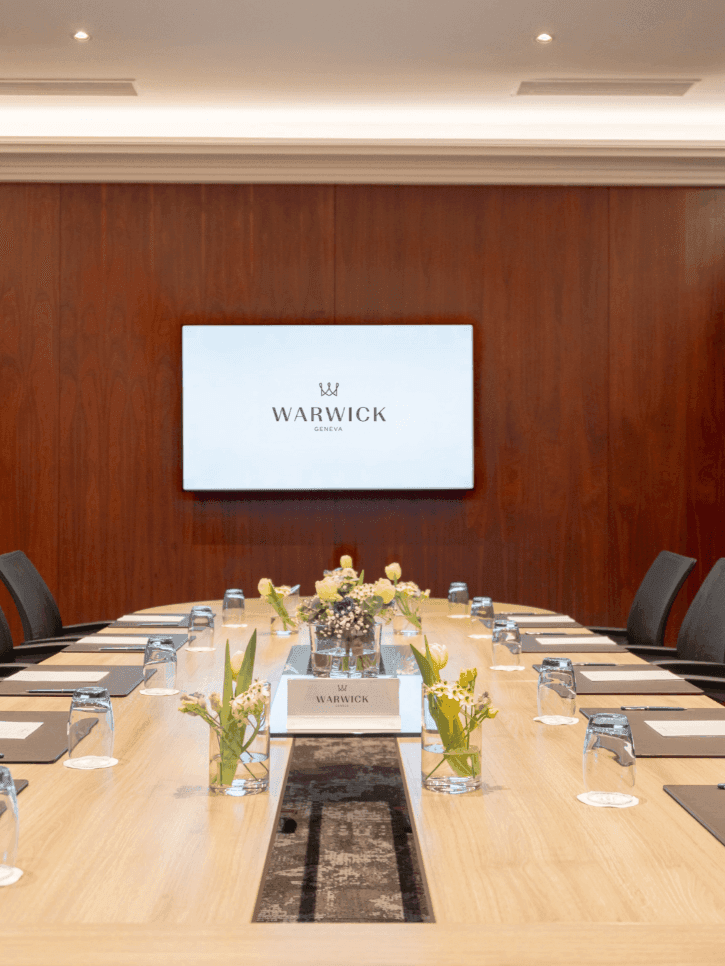 Long oval table by black mesh chairs under a wall-mounted screen in Gobelin at Warwick Geneva