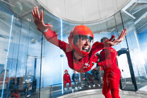 A woman in a red jumpsuit and helmet smiles at the camera, suspended off the ground and being guided by a coach in a red jumpsuit.