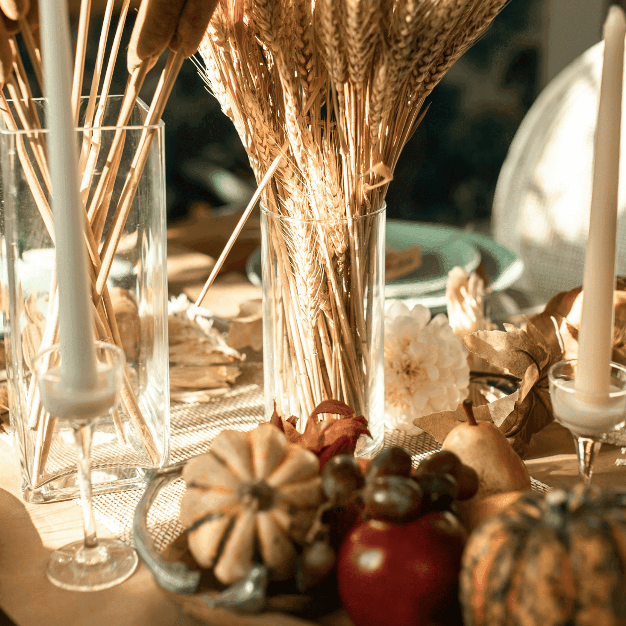 A table with autumnal decor including wheat, pumpkins, and candles in clear glass holders.