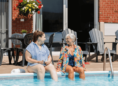 Two smiling women sitting side by side with feet dipped into the clear water of an outdoor hotel pool at Rodd Moncton