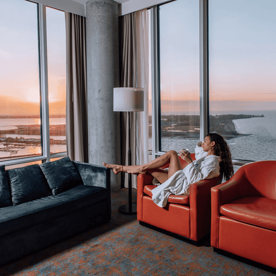 Woman in a robe relaxing on an orange chair beside a navy sofa, overlooking the waterfront at sunset at Hotel X Toronto