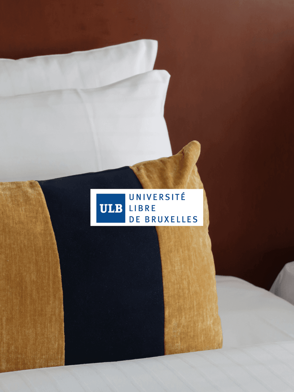 Bed met witte kussens en goudkleurig sierkussen met ULB-logo tegen een terracotta muur