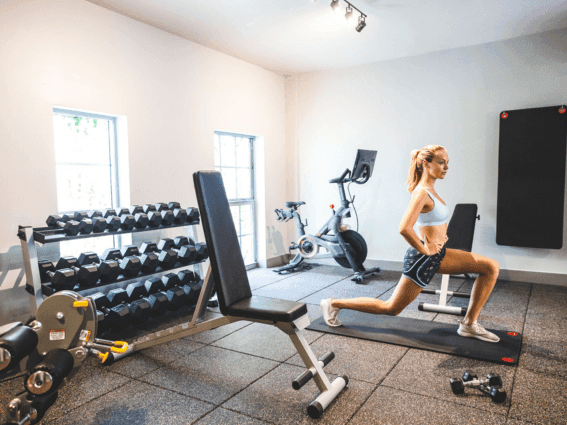 Woman performs a lunge exercise in a bright gym with free weights and a workout bench at The Plymouth Hotel South Beach